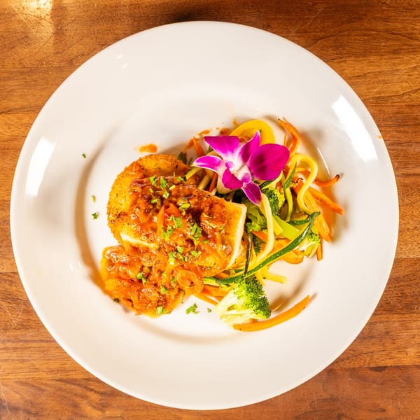 Crispy fried fish with sauce, colorful vegetable noodles, and a purple orchid on a white plate.
