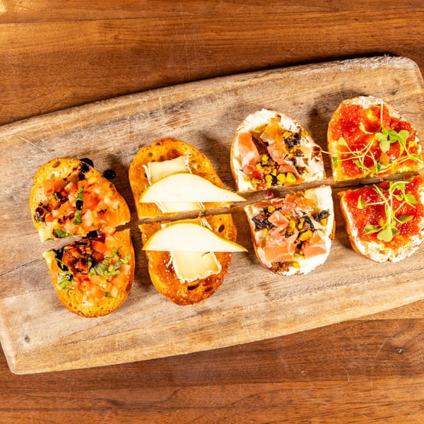 Four varieties of bruschetta, including tomato, pear and brie, prosciutto, and red pepper, on a wooden board.