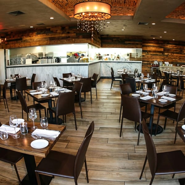 Spacious restaurant dining room with set tables, an open kitchen, and warm brick and wood decor.