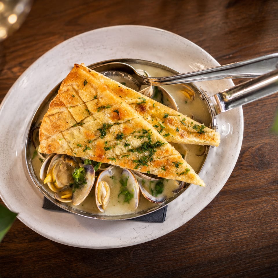 Golden herb-topped flatbread served over open-shell clams in creamy broth on a white plate.