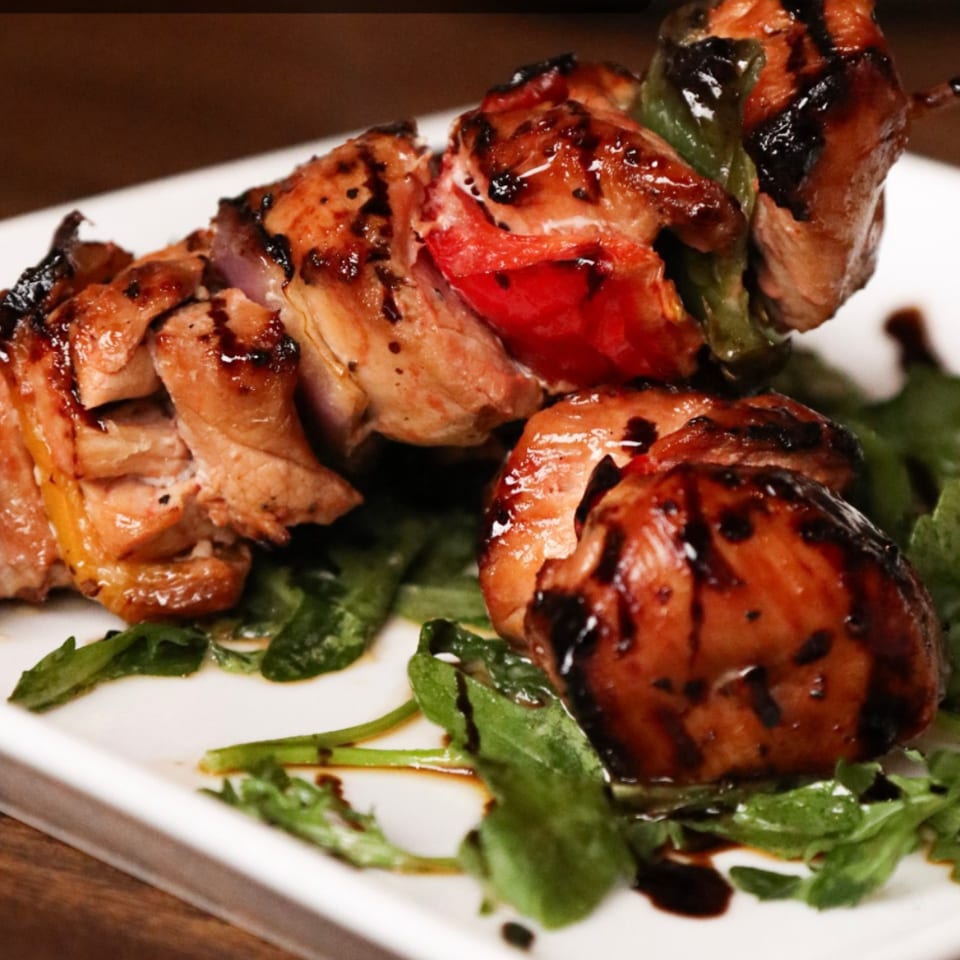 Grilled meat and vegetables threaded onto skewers, served over fresh greens with a balsamic drizzle on a white plate.
