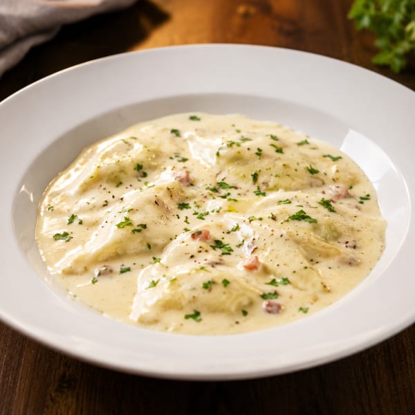Creamy ravioli garnished with parsley and black pepper, served in a white bowl on a wooden table.