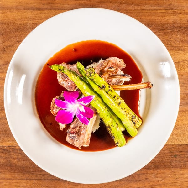 Grilled lamb chops with asparagus, mashed potatoes, and a vibrant purple orchid garnish on a white plate.