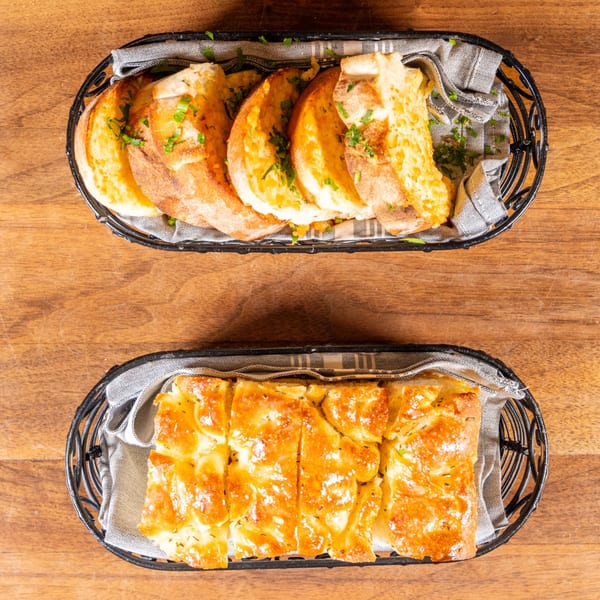 Two baskets of freshly baked bread, one with garlic bread slices and the other with cheesy bread.