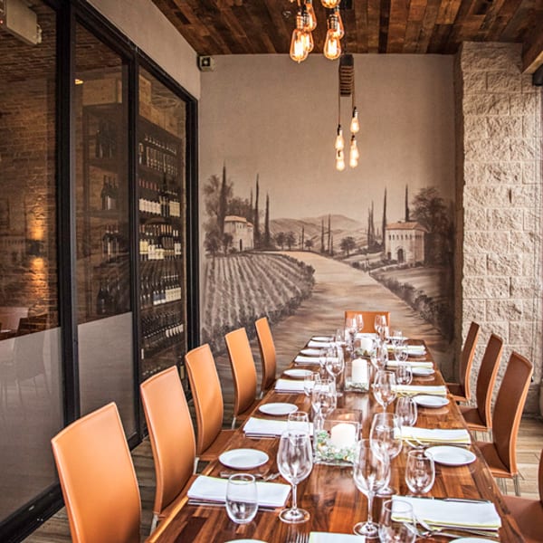Restaurant interior with a long set table, vineyard mural, wine display, and Edison bulb lighting.