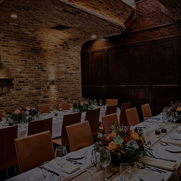 Warmly lit private dining room with long tables set for guests, featuring brick walls and floral decor.